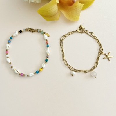 Duas pulseiras femininas, uma colorida com pérolas e outra dourada com estrela do mar e pérolas, sobre fundo claro com flores ao lado.
