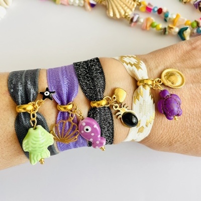 Pulseiras elásticas coloridas com pendentes dourados em pulso, fundo com colares coloridos e conchas