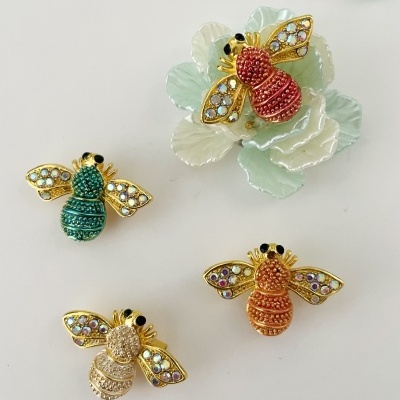 Broches de abelhas douradas com pedras coloridas em quatro cores diferentes, sobre fundo branco com flor cerâmica