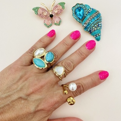 Mão com anéis dourados e unhas pintadas de rosa, broche borboleta rosa e verde, broche concha azul brilhante