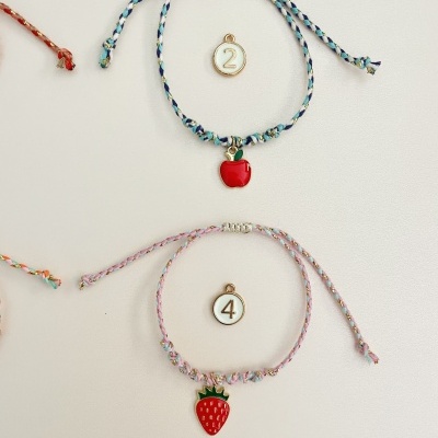 Duas pulseiras trançadas com pendentes de fruta, maçã e morango, e discos numerados 2 e 4.
