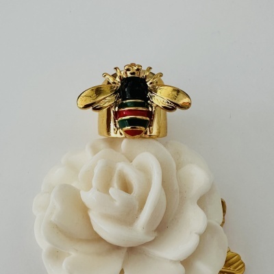 Anel dourado com abelha colorida em esmalte sobre flor branca.