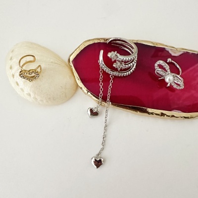 Três anéis decorativos em metal com pedras e pérola sobre fundo branco, concha e base vermelha com borda dourada.