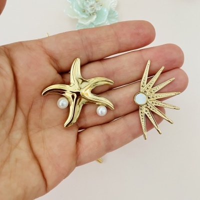 Broches dourados com pérolas em forma de estrela do mar e estrela pontiaguda numa mão com fundo neutro e flores.