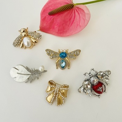 Cinco broches coloridos com detalhes de pérolas e pedras ao lado de uma flor rosa