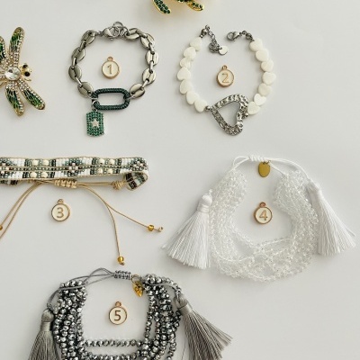 Cinco pulseiras numeradas em diferentes estilos e materiais, com broches decorativos ao fundo, sobre fundo branco.