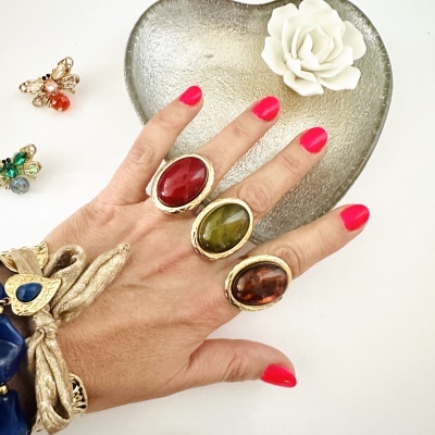 Mão com três anéis grandes e pulseira dourada, coração metálico e flor branca ao fundo