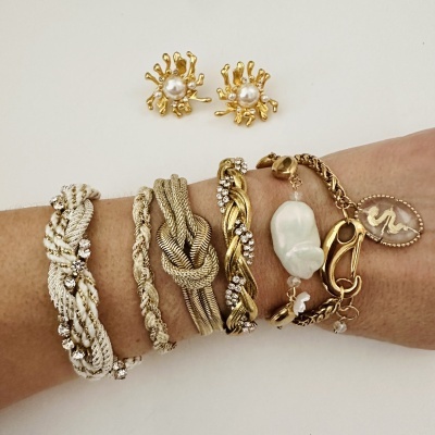 Pulseiras trançadas douradas e prateadas com pedras e pérolas no braço e brincos dourados com pérola ao fundo