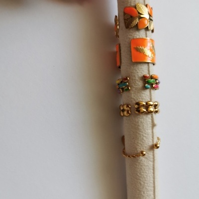 Expositor de veludo bege com seis anéis dourados com detalhes coloridos e pedras, fundo branco.