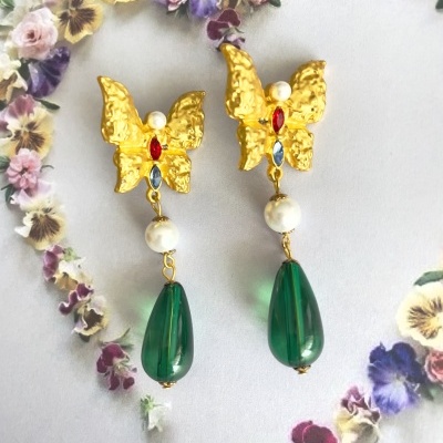 Brincos dourados em forma de borboleta com pedras coloridas, pérola e pendente verde sobre fundo floral.