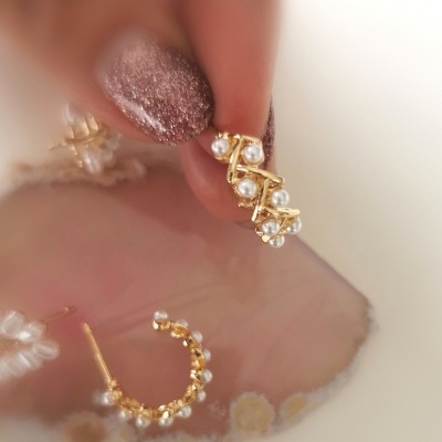 Brincos dourados com pérolas falsas em argola segurados pela mão com unhas rosa brilhante