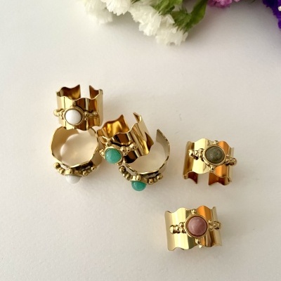 Conjunto de cinco anéis dourados com pedras coloridas e design ondulado sobre fundo branco com flores coloridas
