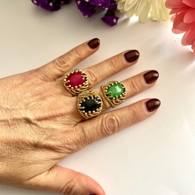 Mão com três anéis grandes de pedras coloridas emolduradas por ouro, com flores ao fundo.
