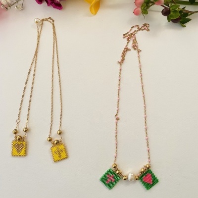 Dois colares delicados com pendentes coloridos e corrente dourada sobre fundo branco e flores coloridas.