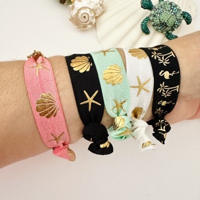 Pulseiras decorativas em tecido com motivos marinhos dourados amarradas num pulso e conchas decorativas ao lado.