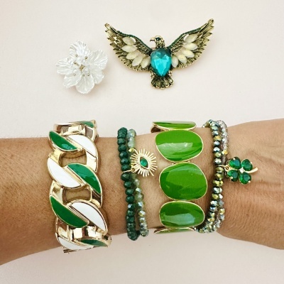 Braço com várias pulseiras verdes e douradas e dois broches decorativos em fundo branco.