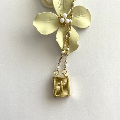 Colar dourado com pendente em forma de livro com cruz e contas brancas sobre flor artificial
