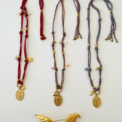 Três colares com medalhões dourados e fios trançados coloridos junto a um broche dourado em forma de pássaro.