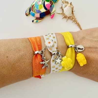 Três braceletes coloridos com pingentes variados mostrados no pulso, ao lado de conchas decorativas. 