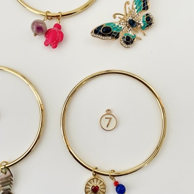 Pulseiras douradas com pendentes coloridos e uma borboleta decorativa num fundo branco.