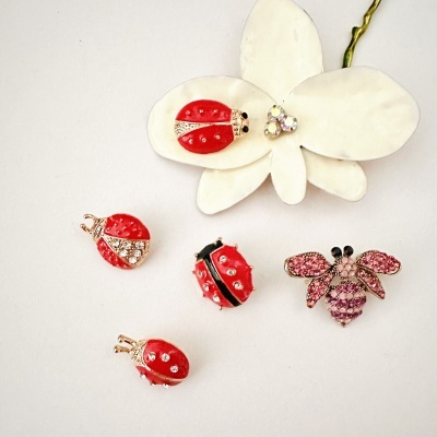 Pins em forma de joaninhas vermelhas e abelha rosa com pedras brilhantes sobre superfície branca com flor artificial