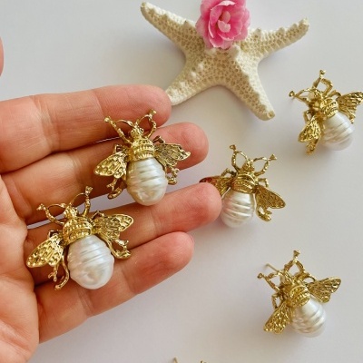 Conjunto de joias em forma de abelha com pérola e metal dourado sobre fundo branco com estrela do mar e flor rosa
