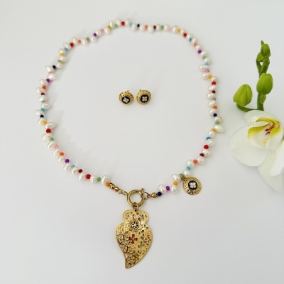 Conjunto de colar e brincos com pérolas e contas coloridas, pendente dourado com cruz vermelha, brincos dourados com detalhes azuis e pérola, flor branca ao lado.