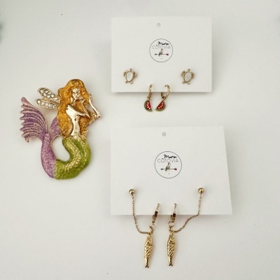 Brincos e broche coloridos de sereia e motivos marinhos em fundo branco.