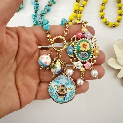 Colar com pendentes decorativos dourados com abelha e flores, segurado por uma mão