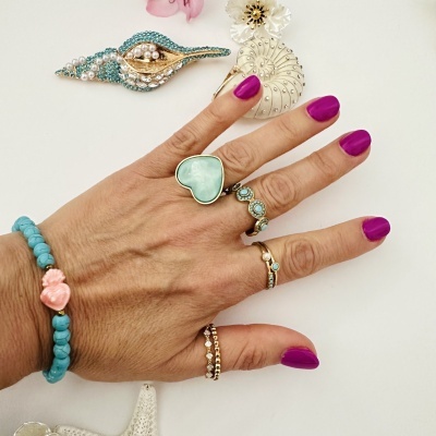 Mão com vários anéis e pulseira, unhas pintadas de roxo, sobre fundo branco com broches decorativos