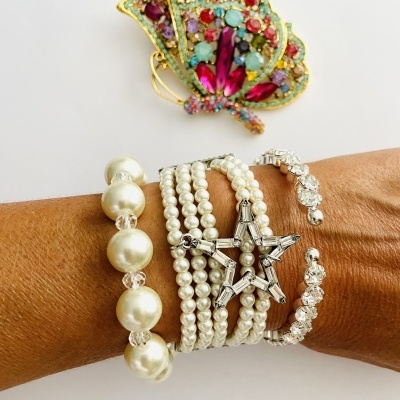 Pulseiras femininas de pérolas e metal prateado com estrela e cristais, e broche colorido ao fundo.