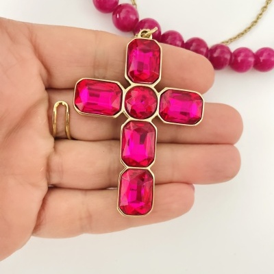 Pendente cruz com pedras fúcsias em metal dourado segurado por mão com anel