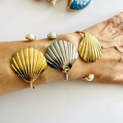 Pulseiras de concha douradas e prateadas com braceletes de pérola em pulso, com conchas naturais e decorativas ao fundo
