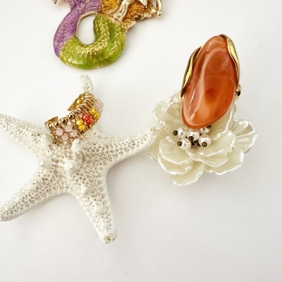 anel dourado com pedras coloridas e broche de sereia colorido sobre fundo branco