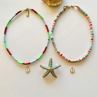 Dois colares coloridos com pendentes numerados, concha dourada com pedras e estrela-do-mar azul sobre fundo branco