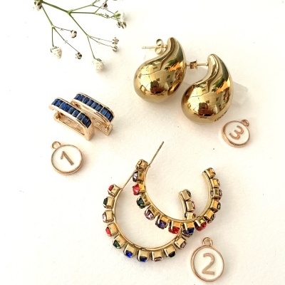 Três pares de brincos dourados com pedras coloridas e números 1, 2 e 3 em círculos brancos sobre fundo branco