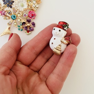 Broche de boneco de neve com chapéu vermelho na mão, vários broches florais ao fundo