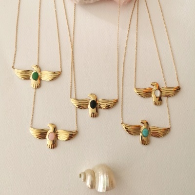 Colares dourados com pendentes de águias e pedras coloridas sobre fundo branco, com uma pedra rosa e uma concha