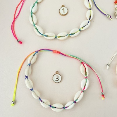 Duas pulseiras de conchas brancas com cordões coloridos numeradas 1 e 3 em fundo claro