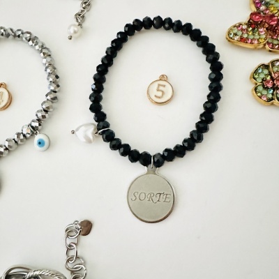 Pulseira preta com pendente prateado escrito SORTE e medalha número 5 num fundo branco.