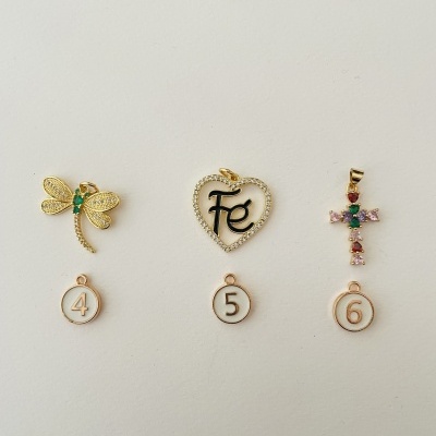 Três pendentes dourados detalhados numerados 4, 5 e 6 com etiquetas incluindo uma libélula, um coração com a palavra Fé, e uma cruz com pedras coloridas