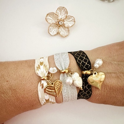 Pulseiras elásticas com pendentes dourados e pérolas num pulso; fundo branco com flor decorativa