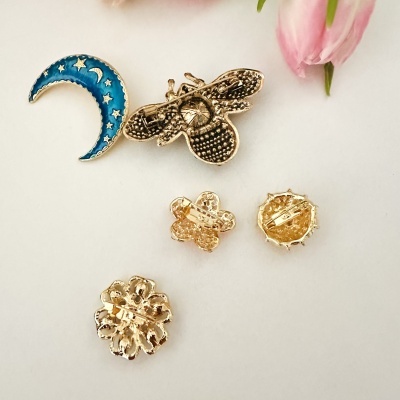 Cinco broches metálicos dourados com formas variadas, incluindo uma lua azul com estrelas, sobre fundo branco e flor rosa.