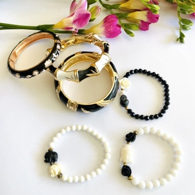 Pulseiras variadas em metal dourado com esmalte preto e pulseiras de contas pretas e brancas com detalhes de caveiras, com flores ao fundo.