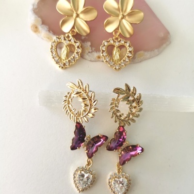 Dois pares de brincos dourados com detalhes em pedra roxa e forma de coração, sobre fundo branco e pedra rosa