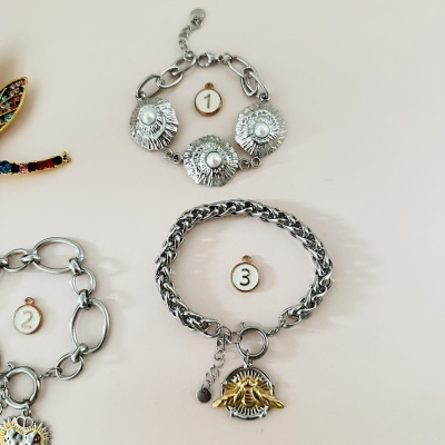 Três pulseiras prateadas numeradas sobre fundo claro, com diferentes designs e adornos metálicos.