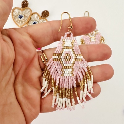 Brinco feito à mão com contas de vidro em rosa, branco e dourado com franjas, segurado por mão com anéis, fundo branco.