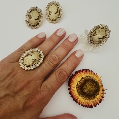 Conjunto de bijuteria com camafeus dourados e pedras brilhantes, mão com anel, unhas nude e flor seca sobre fundo branco