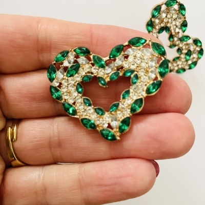 Brincos de coração dourados com pedras verdes e transparentes segurados na mão