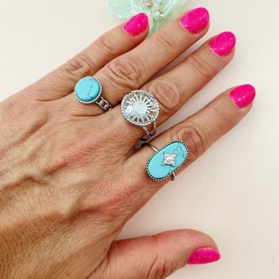 Mão feminina com três anéis azul-turquesa e prateados, unhas cor-de-rosa e flor decorativa branca ao lado.